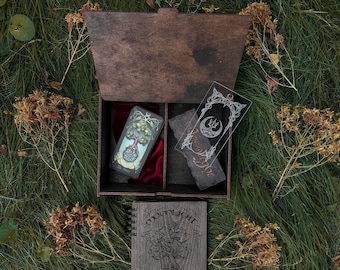 May include: A wooden box with a velvet interior, containing a deck of tarot cards and a clear acrylic bookmark with a moon and star design. The box is open and sitting on a bed of green grass with dried flowers. A wooden journal with the title "Moonlight" is in front of the box.