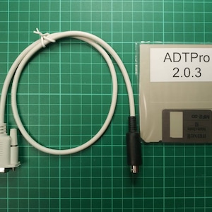 Könnte beinhalten: Ein weißes Kabel mit einem 9-poligen Stecker an einem Ende und einem 5-poligen Stecker am anderen Ende. Eine Diskette mit der Aufschrift "ADTPro 2.0.3" ist ebenfalls im Bild.