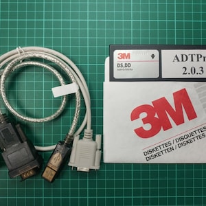 Può includere: Un floppy disk bianco 3M con il testo "ADTPro 2.0.3" e "DISKETTES / DISQUETTES DISKETTEN / DISKETTES" stampato su di esso. Il disco è collegato a un cavo grigio con due connettori.