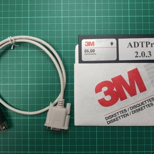 Könnte beinhalten: Eine weiße 3,5-Zoll-Diskette mit der Aufschrift "ADTPro 2.0.3" in einer weißen 3M-Diskettenhülle. Ein weißes Kabel mit einem schwarzen Stecker an einem Ende und einem weißen Stecker am anderen Ende ist ebenfalls abgebildet.