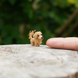 Może przedstawiać: Miniaturowa, szydełkowa figurka psa w odcieniach brązu i beżu. Mały piesek ma puszysty ogon i stoi na jasnej, teksturowanej powierzchni. Tło jest rozmyte, zielone, sugerujące otoczenie na zewnątrz.