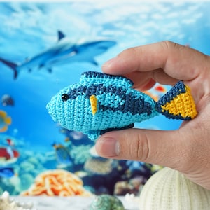 May include: A crocheted blue and yellow fish toy with a black eye. The fish is being held in a hand against a background of a blue and green underwater scene.