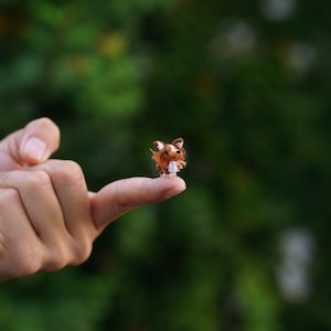 May include: A miniature, handmade crochet dog figurine, primarily brown and white, perched on a fingertip. The tiny dog has detailed features, including pointed ears and a fluffy tail. The background is a blurred green, suggesting an outdoor setting.