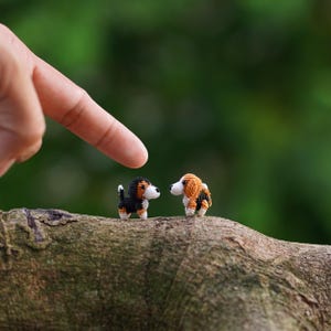Op de afbeelding: Twee kleine gehaakte beagle honden, een zwarte en bruine, de andere oranje en bruin, staan op een tak. Een vinger wijst naar de honden tegen een vage groene achtergrond. De honden hebben witte markeringen en kijken elkaar aan.