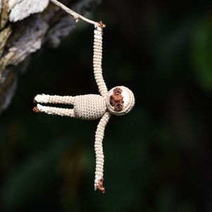 May include: A small crocheted sloth toy hanging from a tree branch. The sloth is beige with brown eyes and a brown nose.