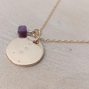 Op de afbeelding: Gouden ketting met een ronde hanger met een sterrenbeeldontwerp en een kleine paarse edelsteenbedel.