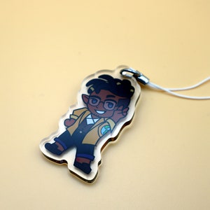 K&ouml;nnte beinhalten: Ein klarer Acryl-Anh&auml;nger mit einer Cartoon-Figur mit dunklem Haar, Brille und gelber Jacke. Der Anh&auml;nger hat einen silbernen Verschluss und ein wei&szlig;es Schl&uuml;sselband. Die Figur l&auml;chelt und zeigt das Friedenszeichen.