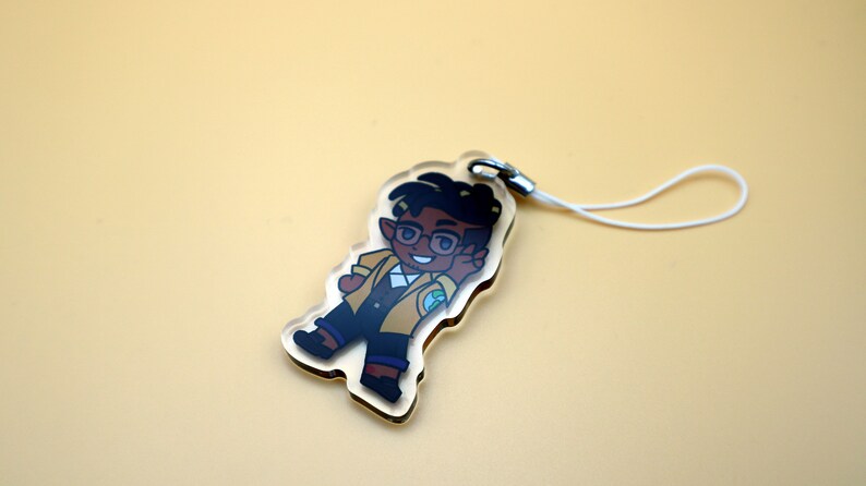 K&ouml;nnte beinhalten: Ein klarer Acryl-Anh&auml;nger mit einer Cartoon-Figur mit dunklem Haar, Brille und gelber Jacke. Der Anh&auml;nger hat einen silbernen Verschluss und ein wei&szlig;es Schl&uuml;sselband. Die Figur l&auml;chelt und zeigt das Friedenszeichen.