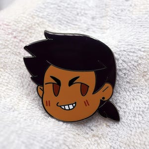 Puede incluir: Un pin que representa la cara de un personaje de dibujos animados con piel morena, pelo negro y ojos marrones. El personaje tiene una expresión traviesa con una pequeña sonrisa y un solo pendiente. El pin está sobre un fondo blanco texturizado.