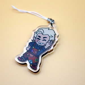 K&ouml;nnte beinhalten: Ein klarer Acryl-Anh&auml;nger mit einer Cartoonfigur mit blonden Haaren, die ein dunkelblaues Hemd und eine braune Latzhose tr&auml;gt. Der Anh&auml;nger hat eine wei&szlig;e Schnur zum Aufh&auml;ngen. Die Figur l&auml;chelt und winkt.