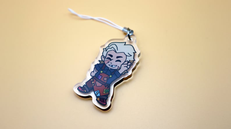 K&ouml;nnte beinhalten: Ein klarer Acryl-Anh&auml;nger mit einer Cartoonfigur mit blonden Haaren, die ein dunkelblaues Hemd und eine braune Latzhose tr&auml;gt. Der Anh&auml;nger hat eine wei&szlig;e Schnur zum Aufh&auml;ngen. Die Figur l&auml;chelt und winkt.