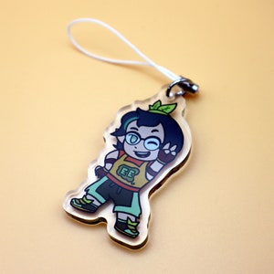 K&ouml;nnte beinhalten: Transparenter Acryl-Anh&auml;nger mit einer Cartoonfigur mit dunklen Haaren, Brille und einem gr&uuml;nen Blatt. Die Figur tr&auml;gt ein gelbes Oberteil mit "EE", gr&uuml;ne Shorts und Schuhe. Ein wei&szlig;es Band ist am Anh&auml;nger befestigt.