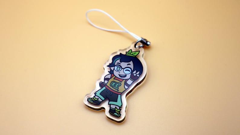 K&ouml;nnte beinhalten: Transparenter Acryl-Anh&auml;nger mit einer Cartoonfigur mit dunklen Haaren, Brille und einem gr&uuml;nen Blatt. Die Figur tr&auml;gt ein gelbes Oberteil mit "EE", gr&uuml;ne Shorts und Schuhe. Ein wei&szlig;es Band ist am Anh&auml;nger befestigt.