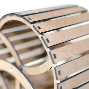 May include: A curved, slatted structure made of light brown composite wood material with dark edges. The object features a series of parallel horizontal slats arranged in a cylindrical arc, connected by thin, dark vertical supports at the ends.
