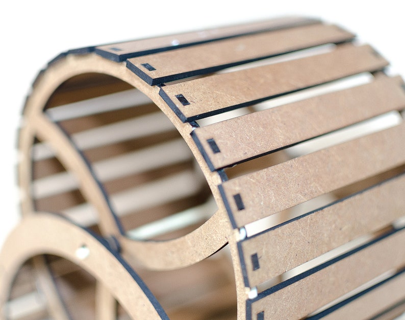May include: A curved, slatted structure made of light brown composite wood material with dark edges. The object features a series of parallel horizontal slats arranged in a cylindrical arc, connected by thin, dark vertical supports at the ends.
