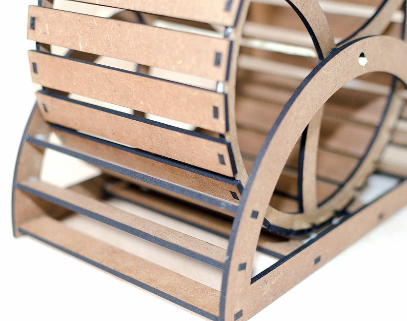 May include: A light brown wooden organizer featuring a slatted, curved design with dark edges. The structure is composed of interlocking panels forming a cylindrical shape with horizontal slats, showcasing a layered, geometric construction.
