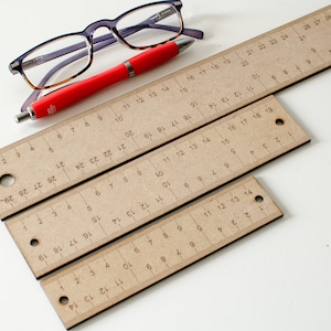 Puede incluir: Tres reglas de madera de diferentes tamaños, con marcas en pulgadas. Las reglas están hechas de cartón marrón y tienen un agujero en el extremo de cada regla. También hay una pluma roja y un par de gafas en la imagen.
