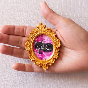 May include: A small, ornate gold-colored frame holds a miniature painting of a sleeping black cat on a pink background. The frame has a detailed, textured design. The artwork is held in a hand.