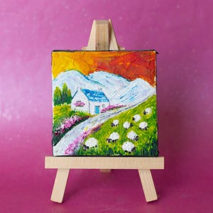 Peut inclure: Petite peinture sur chevalet en bois représentant un paysage avec une maison blanche, un toit bleu et une route sinueuse dans un champ vert avec des moutons. Le ciel est orange et rouge, et les montagnes sont blanches. Des fleurs roses bordent la route.