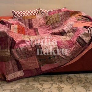 Peut inclure: Une courtepointe patchwork aux tons de rose, de rouge et de marron recouvre un canapé rouge. La courtepointe présente une variété de carrés de tissu cousus ensemble. Deux coussins décoratifs avec un motif géométrique sont visibles derrière la courtepointe. Le texte "studio nakra" est visible.