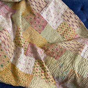 May include: A patchwork quilt made of various shades of beige, pink, and green fabric. The quilt is made of squares of different sizes and is stitched together with a running stitch. The quilt is folded and draped over a blue couch.
