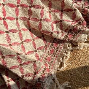 Könnte beinhalten: Ein cremefarbenes Textil mit einem sich wiederholenden Muster aus rosa Blütenformen und braunen Quadraten. Der Rand weist ein florales Design in Rosa und Braun mit Fransen auf. Der Stoff scheint gewebt zu sein.