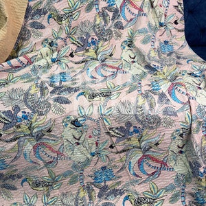 May include: Pink fabric with a repeating pattern of monkeys, birds, and foliage in shades of blue, green, and yellow. The fabric has a textured appearance and is draped over a blue surface.