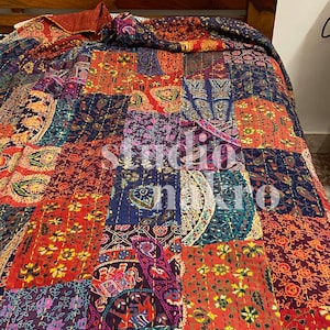 May include: A colourful patchwork quilt with a paisley and floral design. The quilt is made of various fabrics in shades of red, blue, green, and purple. It is folded on a wooden bed frame.