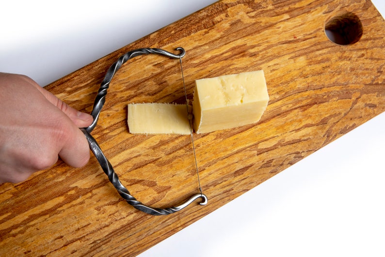 May include: A hand holds a twisted metal cheese wire cutter over a block of cheese on a wooden cutting board. A slice of cheese has been cut from the block.