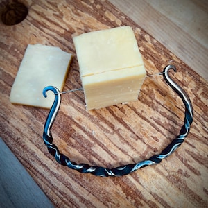 May include: A twisted metal cheese knife with a wire cutter and a wooden chopping board with a block of cheddar cheese.