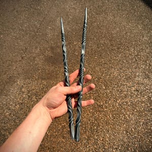 May include: Two hand-forged metal tongs with a twisted design. The tongs are made of dark gray metal and have pointed tips. The handles have a spiral pattern. The tongs are held in a hand against a background of small pebbles.