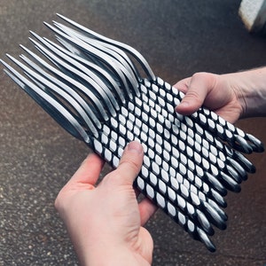 May include: A bundle of 18 silver metal skewers with a black and white diamond pattern. The skewers are twisted and have pointed ends.