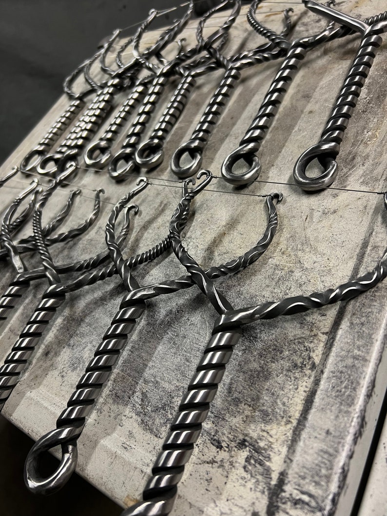 May include: A collection of metal bottle openers with a twisted design. The openers are hanging on a wire rack and have a loop at the top for hanging.