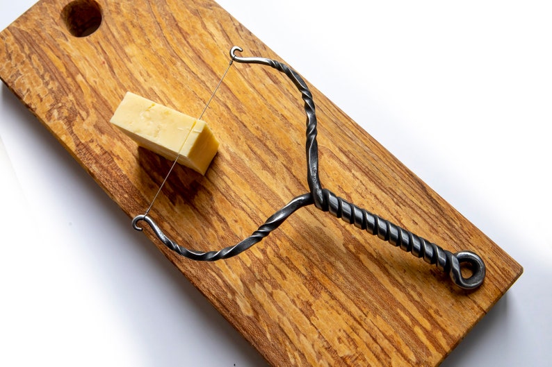 May include: A twisted metal cheese knife with a wire loop on a wooden cutting board with a block of cheddar cheese.