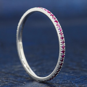 May include: A thin silver band ring featuring a continuous row of small, round, vibrant pinkish-red gemstones set along the outer edge. The metal has a polished, reflective finish, and the stones are evenly spaced, creating a delicate, circular design.