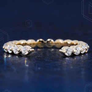 May include: A gold ring with a row of clear, round-cut gemstones. The ring has a split shank design, with the gemstones set along the top half of the band. The ring is displayed on a dark blue surface.