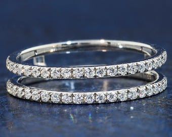 Puede incluir: Un anillo de doble banda en tono plateado con dos filas paralelas de pequeñas piedras transparentes de corte redondo engastadas a lo largo de toda la circunferencia de cada banda. El metal tiene un acabado pulido y brillante que resalta.