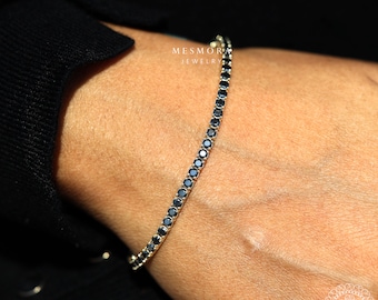 May include: A gold tennis bracelet with a line of small, round, dark blue gemstones. The bracelet is displayed on a person's wrist, with the brand name "MESMORA JEWELRY" visible in the background.