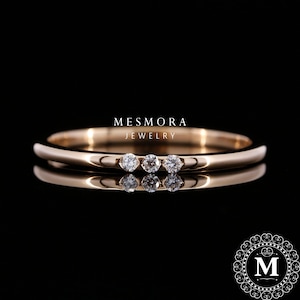 Puede incluir: Un anillo de banda de oro rosa con tres piedras pequeñas, redondas, claras y facetadas colocadas horizontalmente en el centro. El texto MESMORA JEWELRY aparece sobre el anillo y un logotipo circular con la letra M está en la esquina inferior derecha.