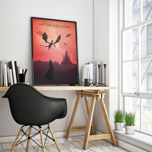 May include: A framed print with a red and orange gradient background. The print features a silhouette of a woman standing in front of a castle with three dragons flying overhead. The text "Mother of Dragons" is at the top of the print.
