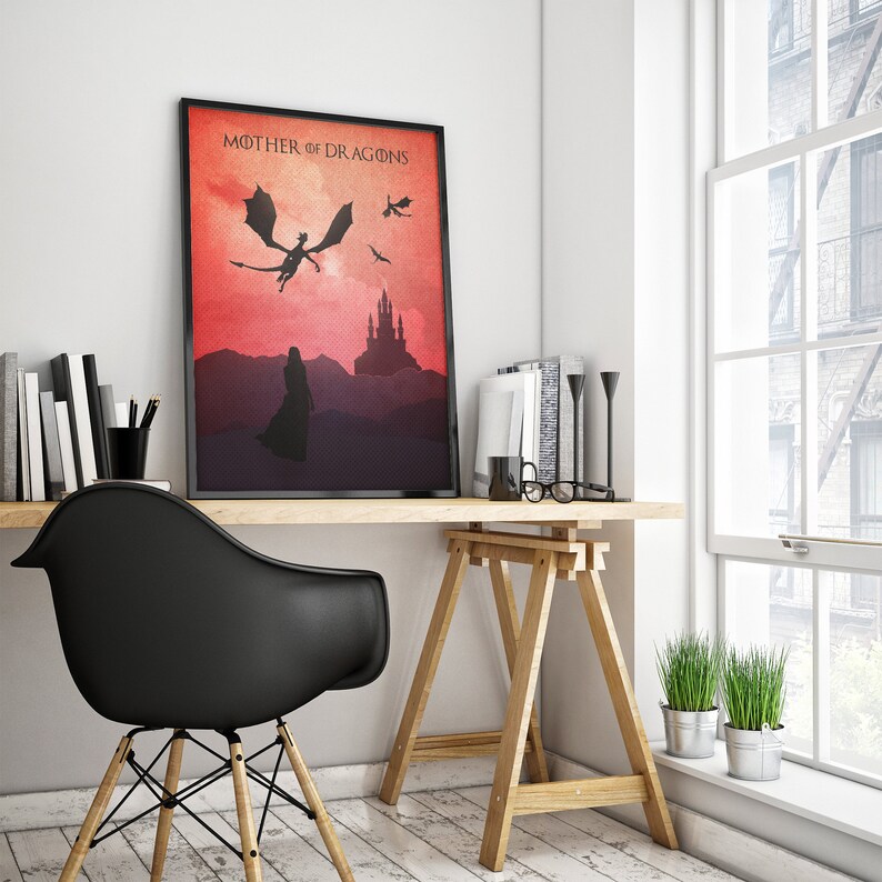 May include: A framed print with a red and orange gradient background. The print features a silhouette of a woman standing in front of a castle with three dragons flying overhead. The text "Mother of Dragons" is at the top of the print.