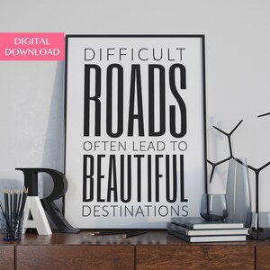 Könnte beinhalten: Schwarz-weißes Typografie-Druck mit dem Zitat "Schwierige Wege führen oft zu schönen Zielen". Der Druck ist in einem schwarzen Rahmen gerahmt und wird auf einem Holzregal mit anderen Dekorationsartikeln ausgestellt.