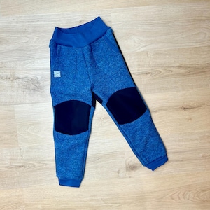 Könnte beinhalten: Blaue Fleecehose mit marinefarbenen Kniepatches. Die Hose hat einen blauen Bund und Bündchen. Auf der linken Seite ist ein kleines Etikett sichtbar. Die Hose ist aus einem weichen, strukturierten Material.