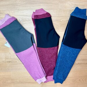 Könnte beinhalten: Drei farbblockierte Leggings in Lila-, Schwarz- und Blautönen. Jedes Paar hat einen einfarbigen Block am oberen Bein und eine kontrastierende Farbe am unteren Bein und am Bund. Die Leggings liegen auf einer hellen Holzoberfläche.