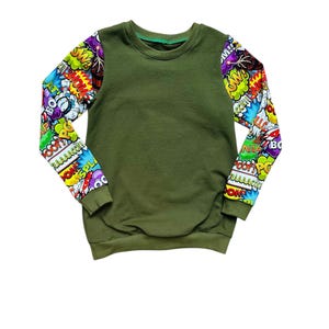 Könnte beinhalten: Olivgrünes Sweatshirt mit langen Ärmeln, die mit einem farbenfrohen Comic-Print versehen sind. Die Ärmel zeigen verschiedene Aktionswörter und Soundeffekte in leuchtenden Farben. Das Sweatshirt mit Rundhalsausschnitt ist ein lässiges Oberteil.