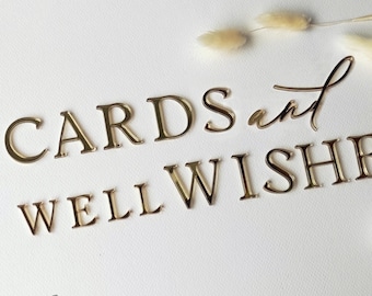 May include: Gold-coloured metal letters spelling "CARDS and WELL WISHES" on a white surface. The word "and" is in a cursive font. Dried flowers are scattered around the text. The Cardcreation Wedding Accessories logo is in the bottom left corner.