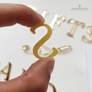 May include: A gold-coloured metal letter "S" is held between fingers, with other gold letters in the background. The letter is in focus, with a slight texture visible. The image is well-lit, highlighting the metallic sheen. The background includes the text "CARDCREATION WEDDING ACCESSORIES".