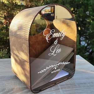Può includere: Una scatola portacarte in legno con parte superiore curva e pannello frontale in acrilico trasparente. L'acrilico presenta la scritta bianca Emily & Leo, un motivo decorativo a tralci con un piccolo cuore e la data 16/06/2026. Contiene buste marroni.
