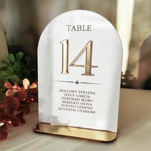 May include: An arched white acrylic table sign featuring the gold-colored text TABLE 14 and a list of six names printed in black. The sign is supported by a rectangular gold-colored base and includes a small decorative horizontal line element.