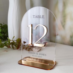 Op de afbeelding: Een boogvormig tafelnummerbord van helder acryl met het nummer 12 in een metallic roségouden afwerking. Het bord bevat de tekst TABLE, de namen OLIVIA & JAMES en de datum 27-07-2027 in het zwart, op een bijpassende roségouden voet.